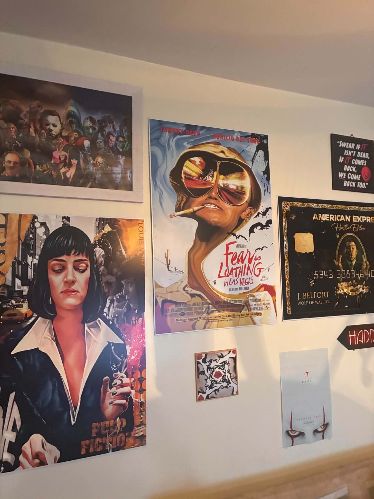 Photo of a real collector's wall covered with framed pop-culture prints: Pulp Fiction, Fear and Loathing in Las Vegas, a horror-icons collage, the Joker, IT, and a gold American Express art card